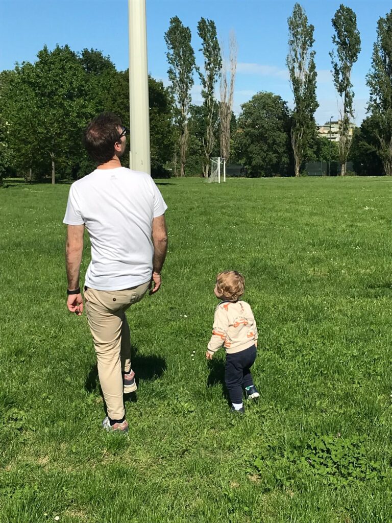 Noia e tempo condiviso: un padre e un bambino camminano insieme su un prato, ripresi a distanza durante una giornata di sole.