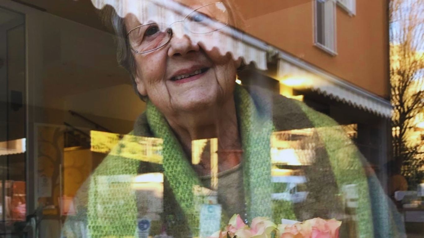 Vivere in RSA: mamma sorridente dietro una vetrata, con un mazzo di rose rosa.